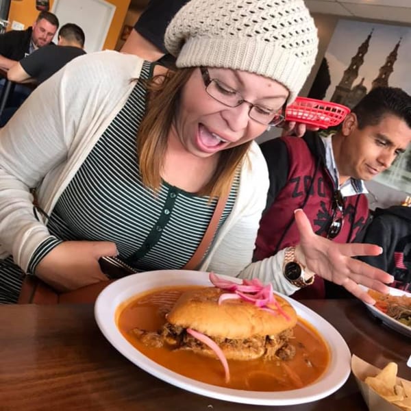 A woman with an excited expression looks at a large Mexican torta in a bowl of red sauce.