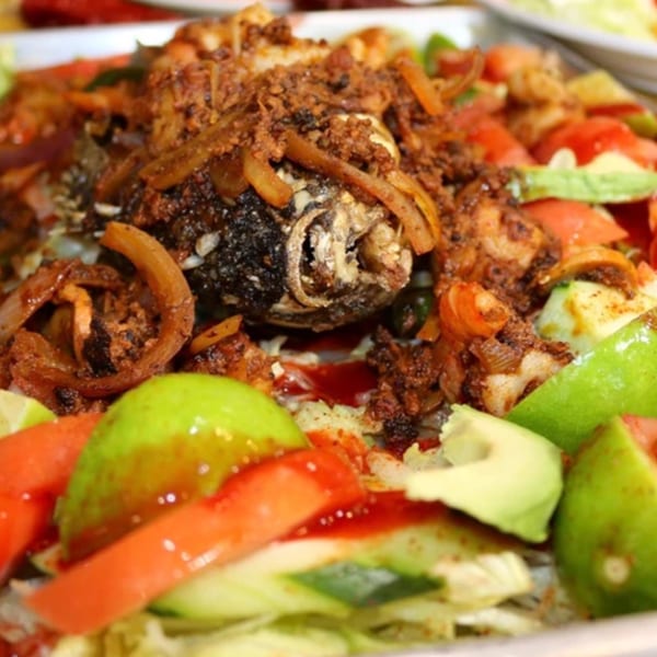Whole fried fish, seasoned with spices and onions, surrounded by fresh avocado, tomato, and lime.