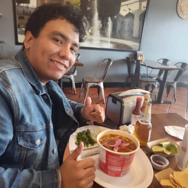 A man in a denim jacket gives a thumbs-up next to a bowl of birria at La Parrilla Taqueria.